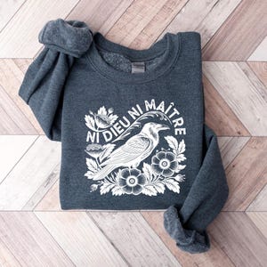 Puede incluir: Una sudadera gris oscuro con un gr&aacute;fico blanco de un cuervo, flores y el texto "NI DIEU NI MA&Icirc;TRE". El dise&ntilde;o est&aacute; centrado en el pecho de la sudadera. Las mangas est&aacute;n enrolladas.