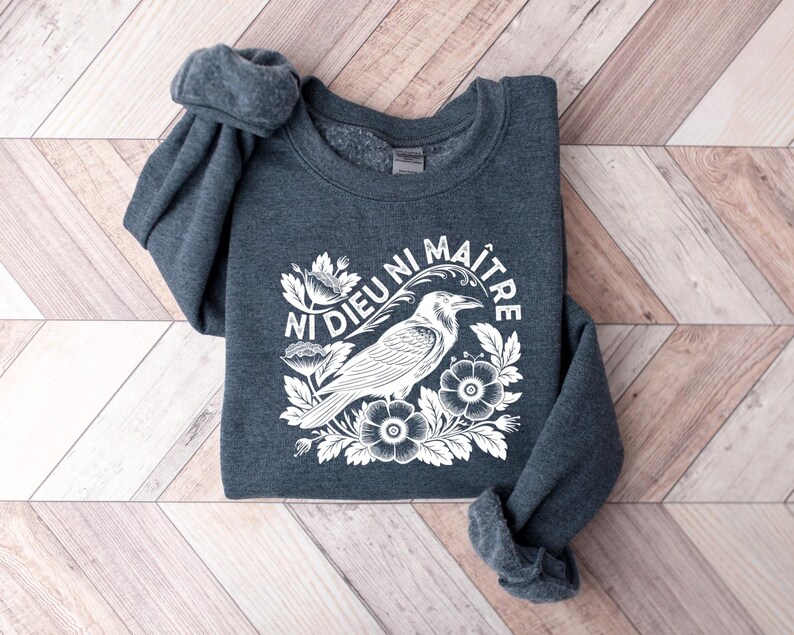 Puede incluir: Una sudadera gris oscuro con un gr&aacute;fico blanco de un cuervo, flores y el texto "NI DIEU NI MA&Icirc;TRE". El dise&ntilde;o est&aacute; centrado en el pecho de la sudadera. Las mangas est&aacute;n enrolladas.