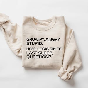 May include: A cream-colored sweatshirt with the text "GRUMPY. ANGRY. STUPID. HOW LONG SINCE LAST SLEEP, QUESTION?" in black, centered on the front. The sleeves are folded up, and the fabric appears soft.