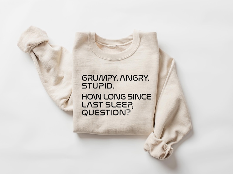 May include: A cream-colored sweatshirt with the text "GRUMPY. ANGRY. STUPID. HOW LONG SINCE LAST SLEEP, QUESTION?" in black, centered on the front. The sleeves are folded up, and the fabric appears soft.