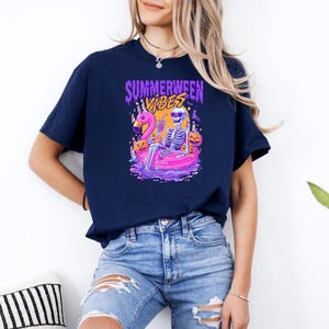 K&ouml;nnte beinhalten: Marineblaues T-Shirt mit einer farbenfrohen Grafik. Die Grafik zeigt ein Skelett auf einem rosa Flamingo-Schwimmer mit dem Text "Summerween Vibes". Das Design beinhaltet K&uuml;rbisse und einen Vollmond.