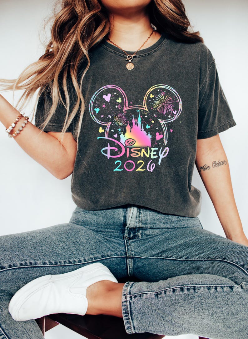 May include: Dark gray t-shirt featuring a colorful Disney 2020 graphic. The design includes a castle, fireworks, and Mickey Mouse ears. The shirt is paired with blue jeans and white sneakers.