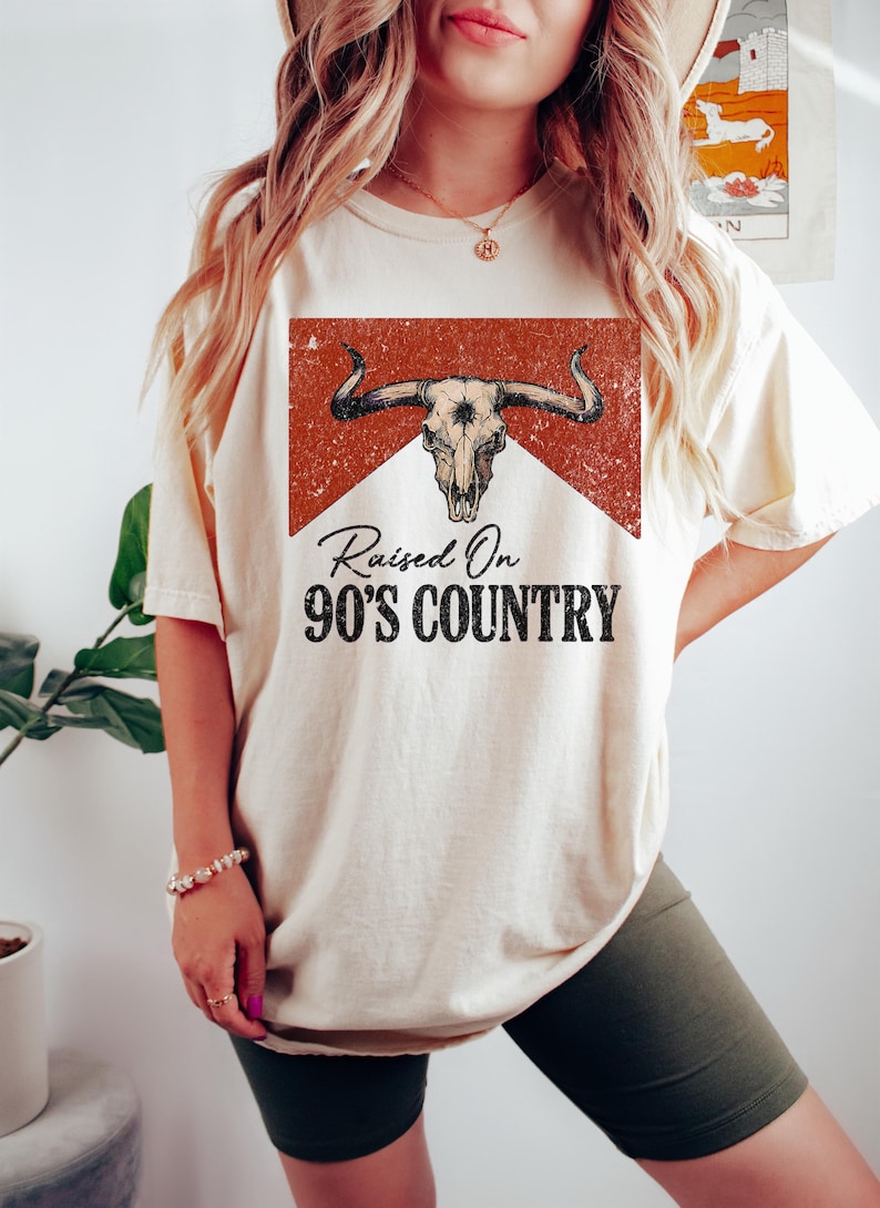 May include: A cream-coloured t-shirt with a distressed bull skull graphic and the text "Raised On 90's Country" in black. The design is set against a red and white background, giving a vintage feel. This is a casual wear item.