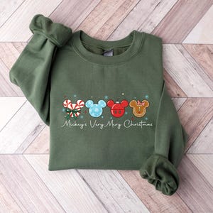 May include: An olive green sweatshirt featuring a festive design with four Mickey Mouse head silhouettes. Each silhouette is decorated with Christmas-themed elements like candy canes, snowflakes, and gingerbread. The text "Mickey's Very Merry Christmas" is written below the design.