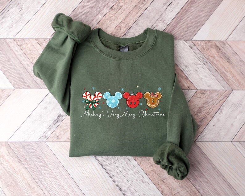 May include: An olive green sweatshirt featuring a festive design with four Mickey Mouse head silhouettes. Each silhouette is decorated with Christmas-themed elements like candy canes, snowflakes, and gingerbread. The text "Mickey's Very Merry Christmas" is written below the design.
