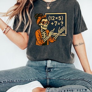 May include: Dark grey t-shirt with a skeleton teacher graphic, wearing glasses and a headband, holding a pointer. The skeleton is in an orange jacket, and the blackboard shows a maths problem: (12 x 5) + 7 = ?. The shirt is worn with blue jeans.