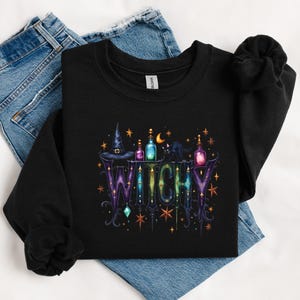May include: Black sweatshirt with a colorful "Witchy" design. The graphic includes a witch hat, potion bottles, a black cat, and stars. The word "Witchy" is in a decorative font with purple and green hues.
