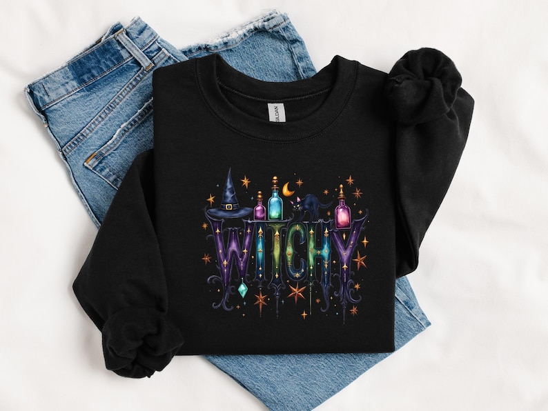 May include: Black sweatshirt with a colorful "Witchy" design. The graphic includes a witch hat, potion bottles, a black cat, and stars. The word "Witchy" is in a decorative font with purple and green hues.