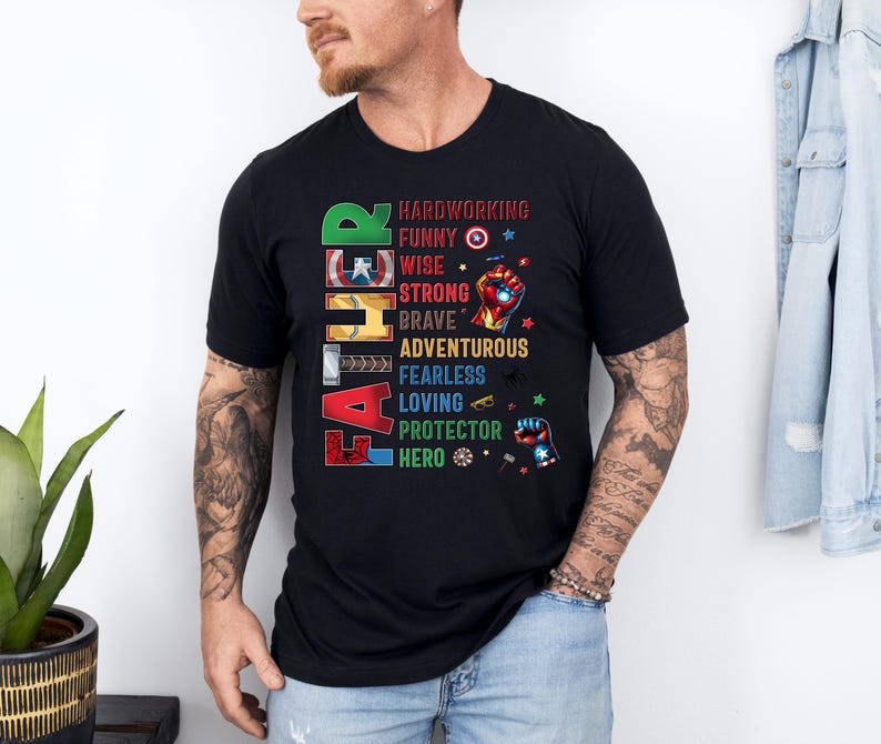 May include: Black t-shirt with the word "FATHER" in colorful block letters. The shirt lists qualities like "hardworking," "funny," and "hero" with superhero-themed graphics. A man is wearing the shirt.