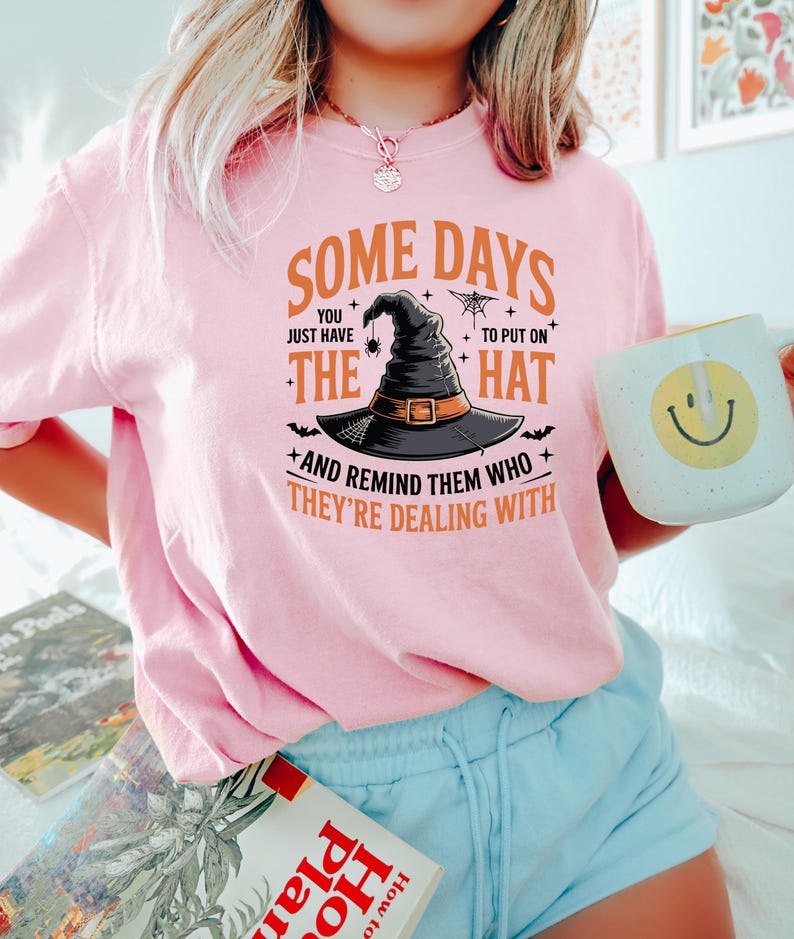 May include: A light pink t-shirt with the text "SOME DAYS YOU JUST HAVE TO PUT ON THE HAT AND REMIND THEM WHO THEY'RE DEALING WITH." The graphic features a witch's hat, spider, and bats. The person is holding a white mug with a yellow smiley face.