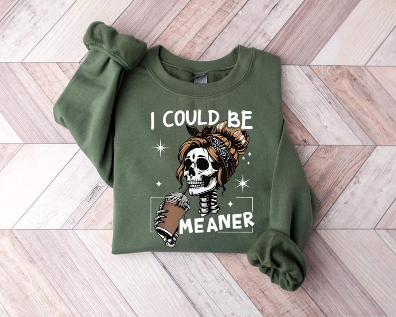 May include: Olive green sweatshirt with the text "I COULD BE MEANER" and a graphic of a skeleton with a bandana drinking from a coffee cup. The design includes star accents and a white rectangular border.