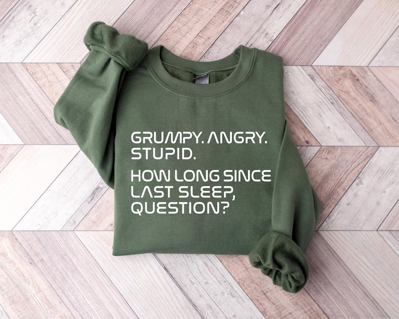 May include: An olive green sweatshirt with the text "GRUMPY. ANGRY. STUPID. HOW LONG SINCE LAST SLEEP, QUESTION?" in white. The sweatshirt is folded on a light brown wooden surface.