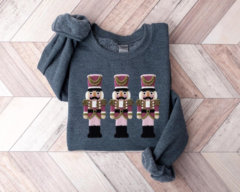 May include: A dark gray sweatshirt featuring an embroidered design of three toy nutcracker soldiers. The nutcrackers have pink and gold accents, black boots, and detailed uniforms. The sweatshirt is folded on a light wood-grain surface.
