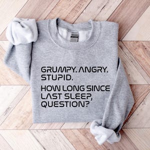 May include: A heather gray sweatshirt with the text "GRUMPY. ANGRY. STUPID. HOW LONG SINCE LAST SLEEP, QUESTION?" in black. The sweatshirt is folded on a light brown wooden surface.
