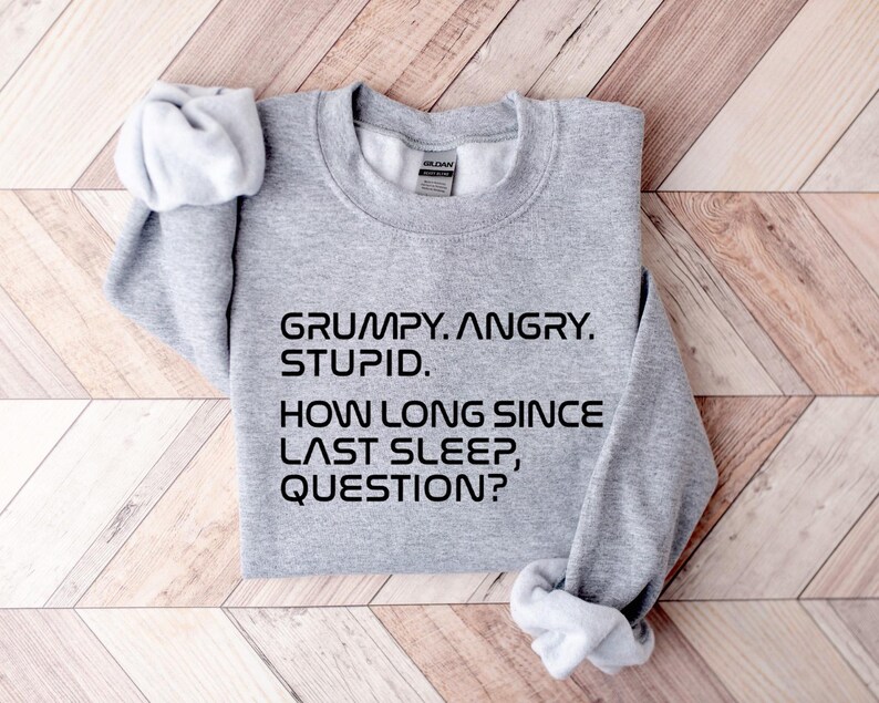 May include: A heather gray sweatshirt with the text "GRUMPY. ANGRY. STUPID. HOW LONG SINCE LAST SLEEP, QUESTION?" in black. The sweatshirt is folded on a light brown wooden surface.
