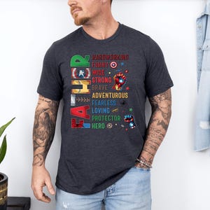 May include: Dark gray t-shirt with the word "FATHER" in colorful block letters. The shirt features a list of positive attributes like "hardworking" and "hero" with superhero-themed graphics. A casual, everyday wear tee.