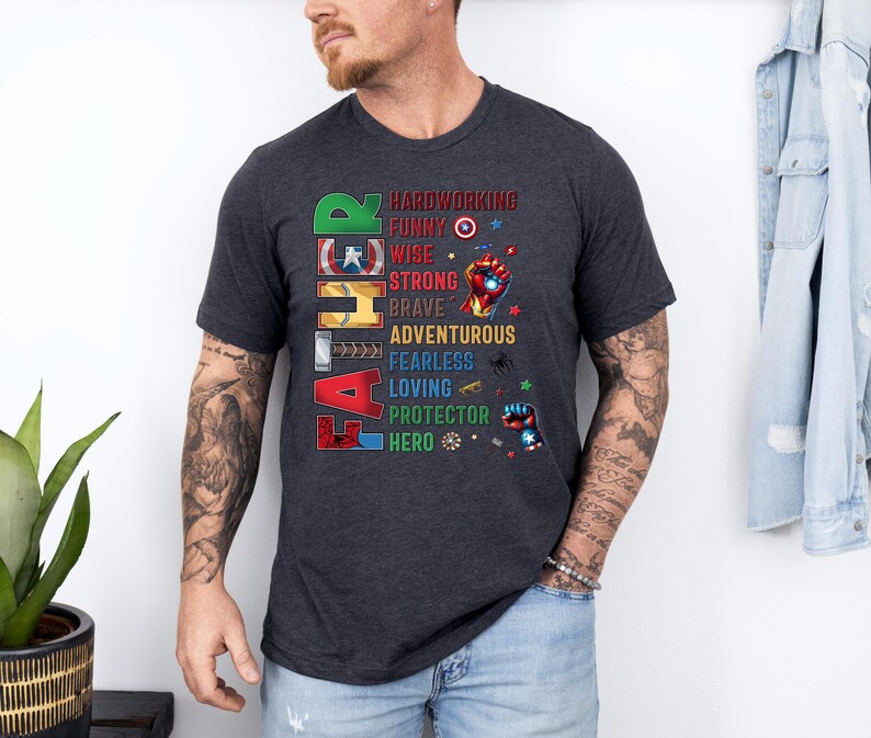 May include: Dark gray t-shirt with the word "FATHER" in colorful block letters. The shirt features a list of positive attributes like "hardworking" and "hero" with superhero-themed graphics. A casual, everyday wear tee.