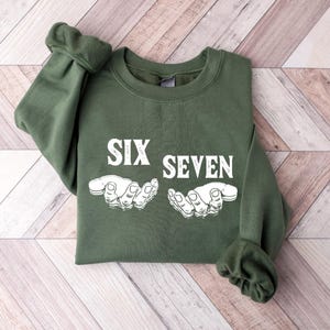May include: An olive green sweatshirt with the words "SIX SEVEN" in white block letters. Below the text are two white illustrations of hands. The sweatshirt is folded on a light wood grain surface.