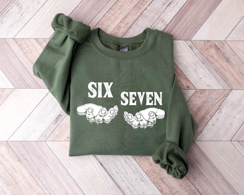 May include: An olive green sweatshirt with the words "SIX SEVEN" in white block letters. Below the text are two white illustrations of hands. The sweatshirt is folded on a light wood grain surface.