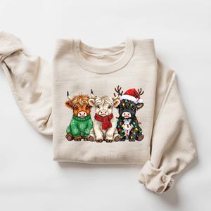 May include: Cream-coloured sweatshirt with a graphic of three Highland cows. The cows are dressed for the holidays: a green sweater, a red scarf, and a Santa hat with fairy lights. A festive and seasonal design.