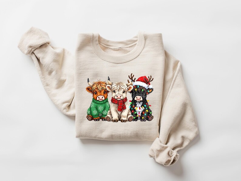 May include: Cream-coloured sweatshirt with a graphic of three Highland cows. The cows are dressed for the holidays: a green sweater, a red scarf, and a Santa hat with fairy lights. A festive and seasonal design.