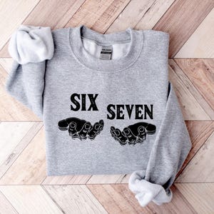 May include: A light gray sweatshirt with the words "SIX SEVEN" in bold black letters. Below the text are two black hand illustrations. The sweatshirt is folded on a wooden surface.