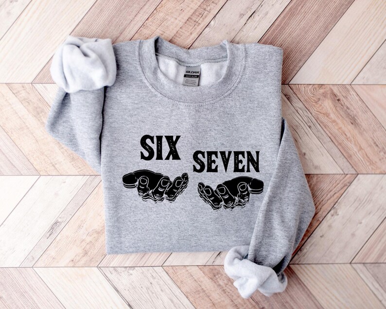 May include: A light gray sweatshirt with the words "SIX SEVEN" in bold black letters. Below the text are two black hand illustrations. The sweatshirt is folded on a wooden surface.