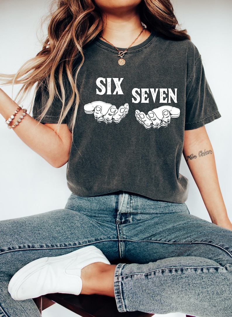 May include: Dark gray t-shirt with the words "SIX SEVEN" in white block letters above two white hand illustrations. The shirt is paired with blue jeans and white slip-on shoes. The overall style is casual and relaxed.