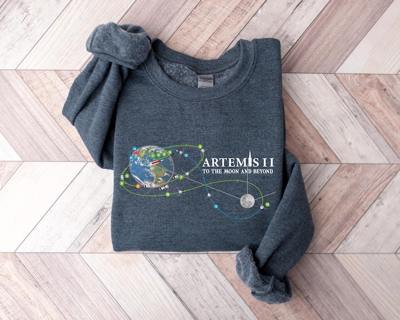 May include: Dark gray sweatshirt with the text "ARTEMIS II TO THE MOON AND BEYOND" and a graphic of Earth, the moon, and connecting lines. The sweatshirt is folded on a light brown and white wooden surface.