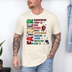 May include: Cream-colored t-shirt with the word "FATHER" vertically down the left side in colorful, comic-book style lettering. The shirt features a list of positive attributes like "hardworking" and "hero" in a matching font.