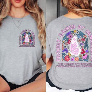May include: A light gray t-shirt with the text "PROUDLY RAISED BY IMMIGRANTS" and a butterfly design. The design includes a butterfly, flowers, and the text "THE CHILDREN OF THOSE WHO TURNED NOTHING INTO SOMETHING".