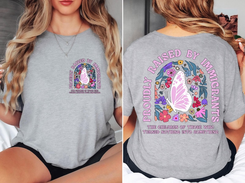 May include: A light gray t-shirt with the text "PROUDLY RAISED BY IMMIGRANTS" and a butterfly design. The design includes a butterfly, flowers, and the text "THE CHILDREN OF THOSE WHO TURNED NOTHING INTO SOMETHING".
