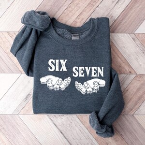 May include: A dark gray sweatshirt with the words "SIX SEVEN" in white block letters and a white graphic of two hands. The sweatshirt is folded on a light brown and white wooden surface.