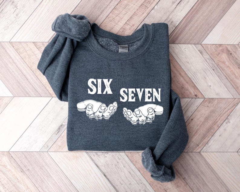 May include: A dark gray sweatshirt with the words "SIX SEVEN" in white block letters and a white graphic of two hands. The sweatshirt is folded on a light brown and white wooden surface.