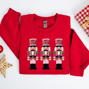 May include: A red sweatshirt featuring three embroidered nutcracker figures in pink, gold, and black. The design is centered on the chest. Christmas-themed decorations, including a wrapped gift and ornaments, are arranged around the sweatshirt.