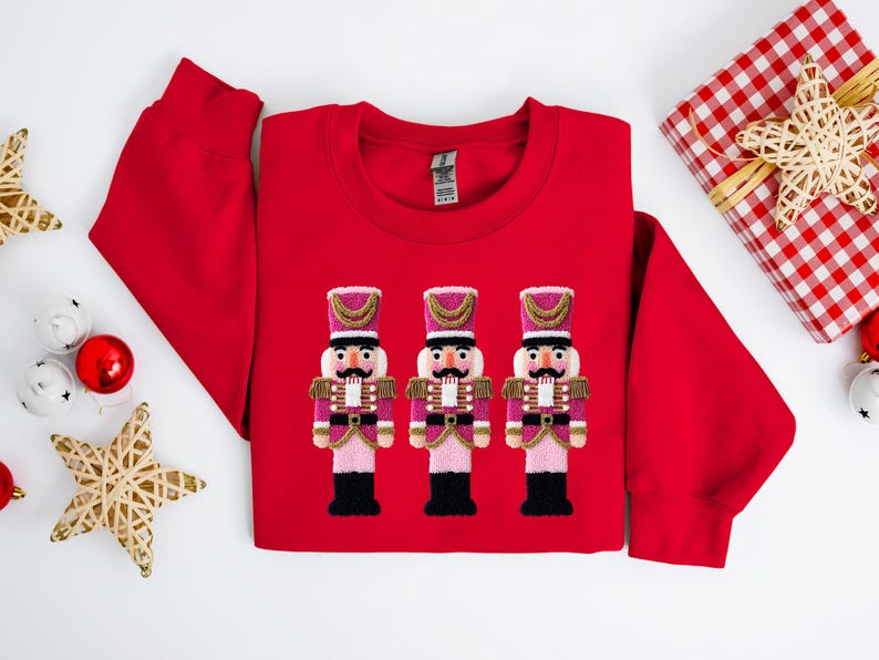 May include: A red sweatshirt featuring three embroidered nutcracker figures in pink, gold, and black. The design is centered on the chest. Christmas-themed decorations, including a wrapped gift and ornaments, are arranged around the sweatshirt.