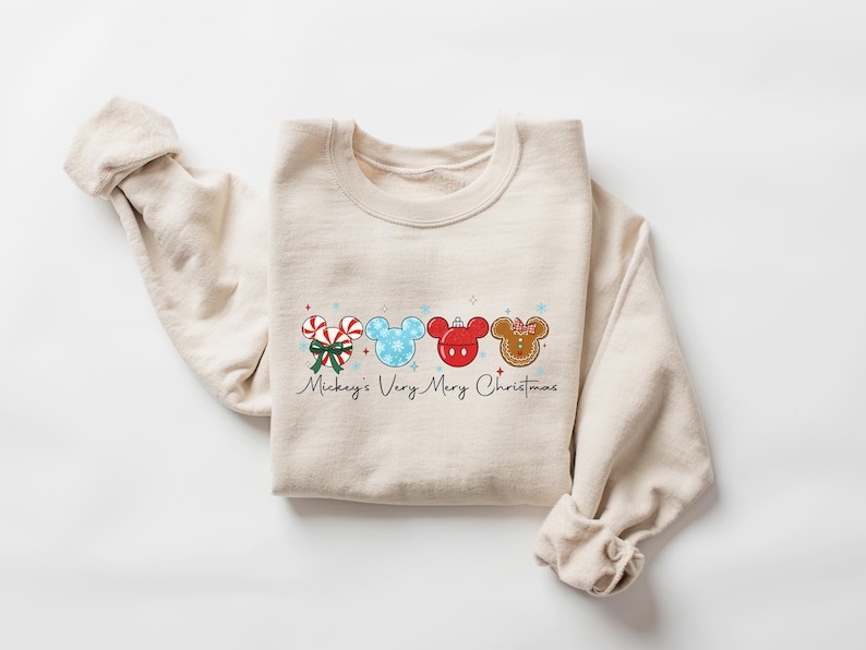 May include: Beige sweatshirt featuring a festive graphic with Mickey Mouse-themed Christmas ornaments. The design includes candy canes, snowflakes, and gingerbread, with the text "Mickey's Very Merry Christmas" in a script font. The sweatshirt is laid flat.