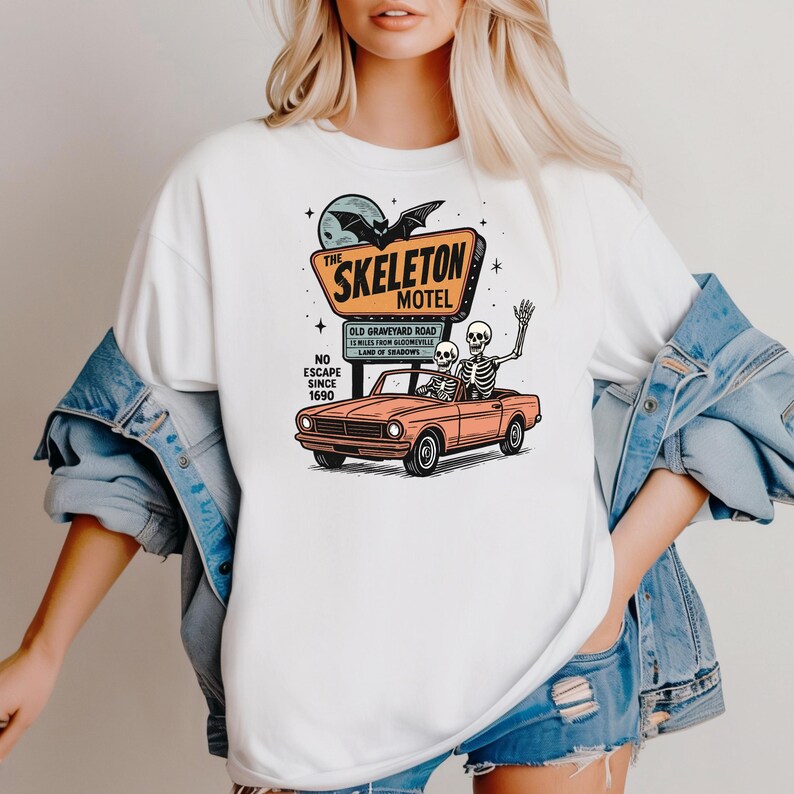 May include: White t-shirt featuring a retro design of "The Skeleton Motel" sign with a car carrying two skeletons. The sign includes a bat, moon, and stars. The car is orange, and the text reads "Old Graveyard Road, 23 miles from Bloomville, Land of Shadows, No Escape Since 1690."