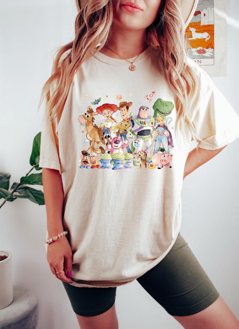 May include: Cream-colored t-shirt featuring a colorful graphic of characters from the animated movie "Toy Story." The design includes Woody, Buzz Lightyear, Jessie, and other beloved characters. The shirt is a casual, comfortable style.