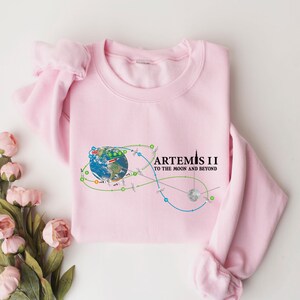 May include: A light pink sweatshirt featuring a graphic of Earth with a trajectory design and the text "ARTEMIS II TO THE MOON AND BEYOND" in black. The design includes green, blue, and orange lines. The sweatshirt is made of a soft material.