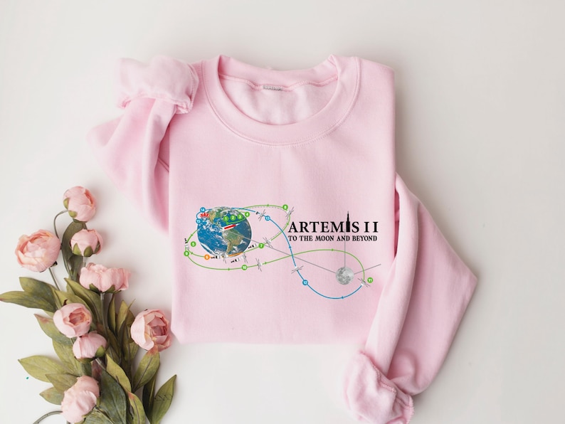 May include: A light pink sweatshirt featuring a graphic of Earth with a trajectory design and the text "ARTEMIS II TO THE MOON AND BEYOND" in black. The design includes green, blue, and orange lines. The sweatshirt is made of a soft material.