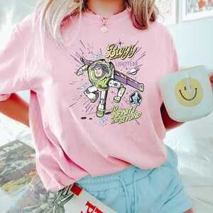 Op de afbeelding: Roze T-shirt met een afbeelding van Buzz Lightyear, met de tekst "Buzz Lightyear" en "To Infinity and Beyond". Het ontwerp toont een cartoonafbeelding van Buzz in zijn ruimtepak. Een witte mok met een gele smiley is ook zichtbaar.