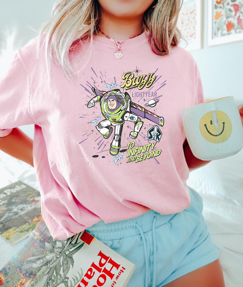 Op de afbeelding: Roze T-shirt met een afbeelding van Buzz Lightyear, met de tekst "Buzz Lightyear" en "To Infinity and Beyond". Het ontwerp toont een cartoonafbeelding van Buzz in zijn ruimtepak. Een witte mok met een gele smiley is ook zichtbaar.