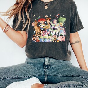 May include: Dark gray t-shirt featuring a colorful graphic of characters from the animated movie Toy Story. The design includes Woody, Buzz Lightyear, Jessie, and other beloved characters. The shirt is made of a soft material.