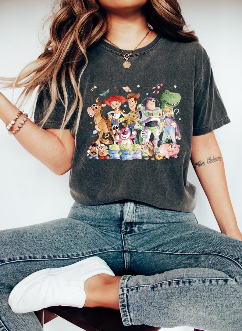 May include: Dark gray t-shirt featuring a colorful graphic of characters from the animated movie Toy Story. The design includes Woody, Buzz Lightyear, Jessie, and other beloved characters. The shirt is made of a soft material.