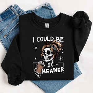 May include: Black sweatshirt with the text "I COULD BE MEANER" and a graphic of a skeleton with a messy bun drinking from a coffee cup. The sweatshirt is laid on top of blue jeans.