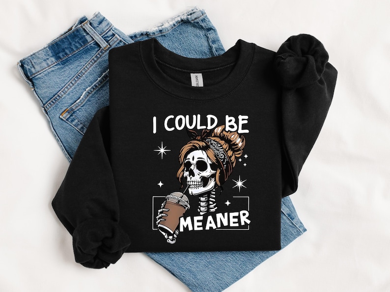 May include: Black sweatshirt with the text "I COULD BE MEANER" and a graphic of a skeleton with a messy bun drinking from a coffee cup. The sweatshirt is laid on top of blue jeans.