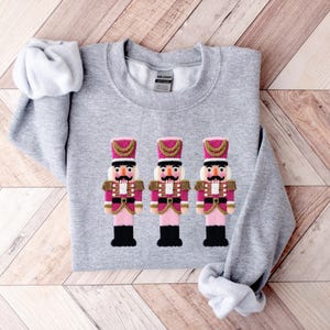 May include: A light gray sweatshirt featuring an embroidered design of three toy nutcracker soldiers. The nutcrackers have pink jackets, black boots, and gold accents. The sweatshirt is made of a soft material.