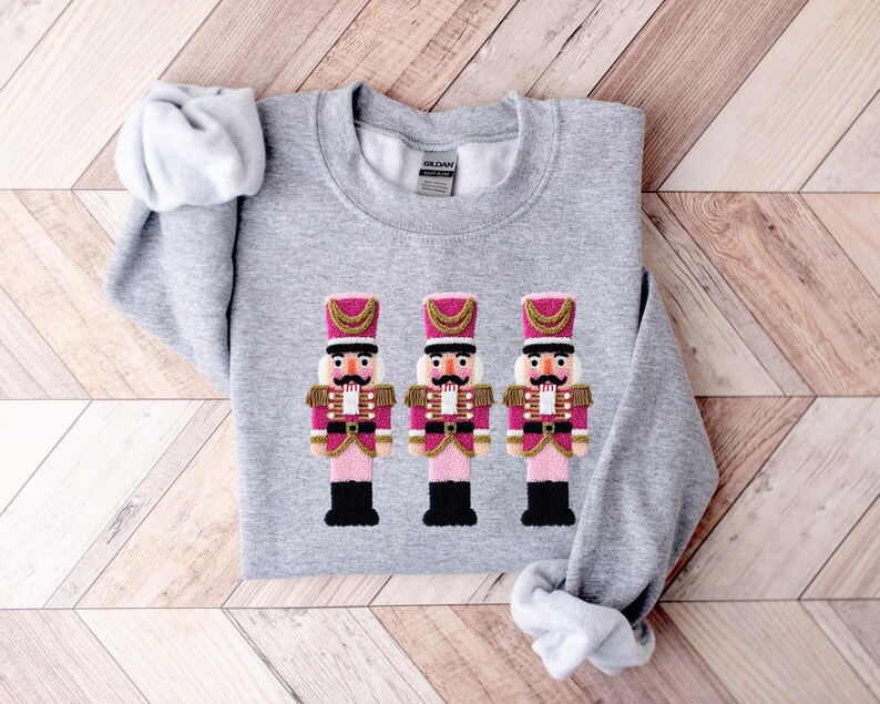 May include: A light gray sweatshirt featuring an embroidered design of three toy nutcracker soldiers. The nutcrackers have pink jackets, black boots, and gold accents. The sweatshirt is made of a soft material.