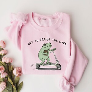 May include: A light pink sweatshirt with the text "OFF TO PRAISE THE LORD" above a cartoon frog riding a scooter while holding a book. The frog is green, and the scooter is white and gray.